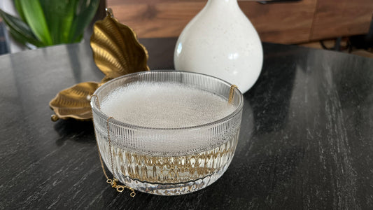 glass bowl with soapy water, gold necklace, marble surface, brass leaf dish, white ceramic vase, elegant decor, sophisticated setting, jewelry cleaning, stylish interior, serene atmosphere.