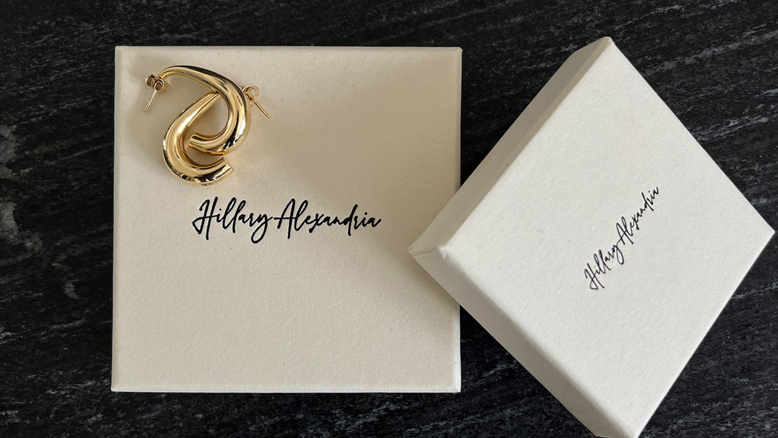 A pair of recycled gold earrings on a cream-colored box with the "Hillary Alexandria" brand logo written in cursive script.