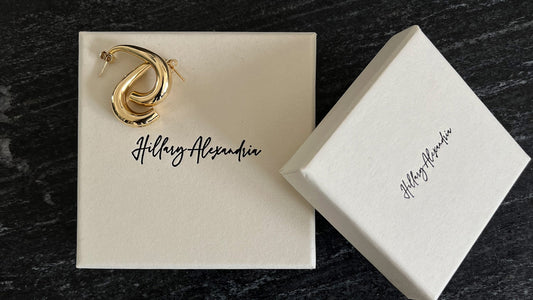 A pair of recycled gold earrings on a cream-colored box with the "Hillary Alexandria" brand logo written in cursive script.